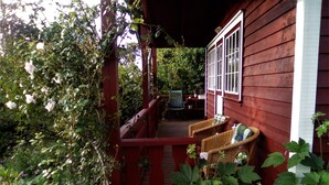 Eine hölzerne Veranda mit Korbstühlen und einer Bank, umgeben von üppigem Grün.