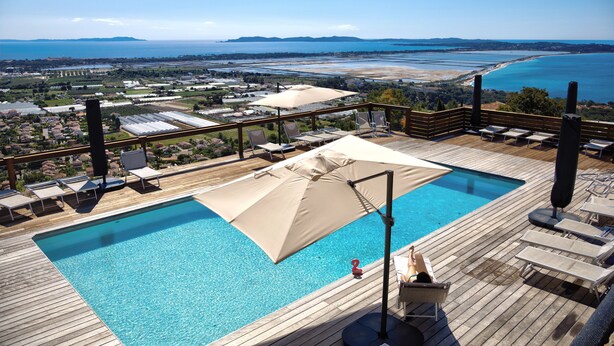 Una piscina con sillas de descanso y sombrillas, rodeada por una terraza de madera y con vistas a un paisaje costero.