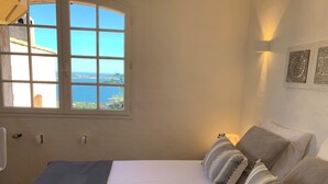 Un dormitorio con un ventanal que ofrece vistas al mar y a edificios. Hay una cama con almohadas a rayas y una mesita de noche.