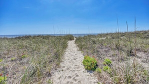 Ein sandiger Pfad führt zum Strand, begleitet von vereinzelter Vegetation und einem wolkenlosen, blauen Himmel.