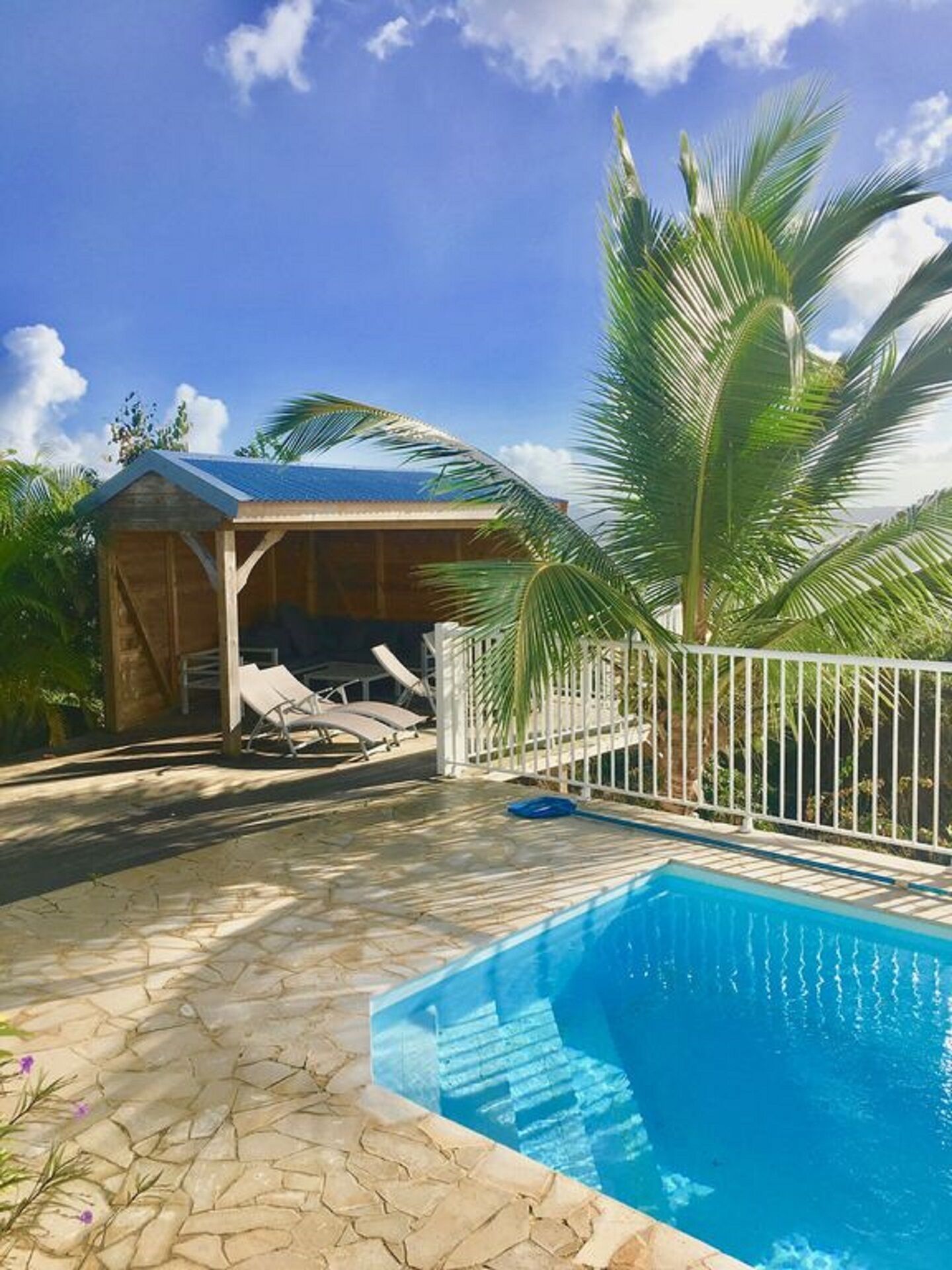 A pool area with a clear blue pool, a covered seating area with lounge chairs, and a wooden structure.