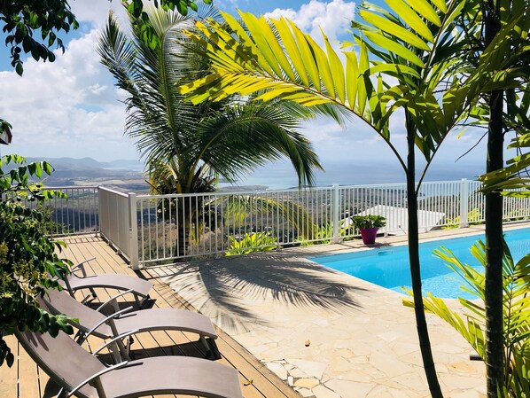 A pool area with lounge chairs, a white fence, and a view of the ocean.