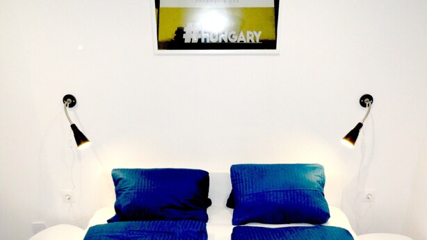 Una cama doble con ropa de cama azul y almohadas blancas, dos lámparas de noche y un cartel enmarcado sobre la cama.