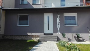Un edificio de dos plantas con fachada gris, una puerta blanca y el nombre 'TRENT' exhibido verticalmente. Hay ventanas en ambos pisos, y un pequeño espacio al aire libre con sillas y mesas.
