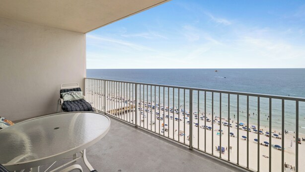 Un balcon avec une table et des chaises, donnant sur une plage parsemée de parasols et fréquentée par des gens.