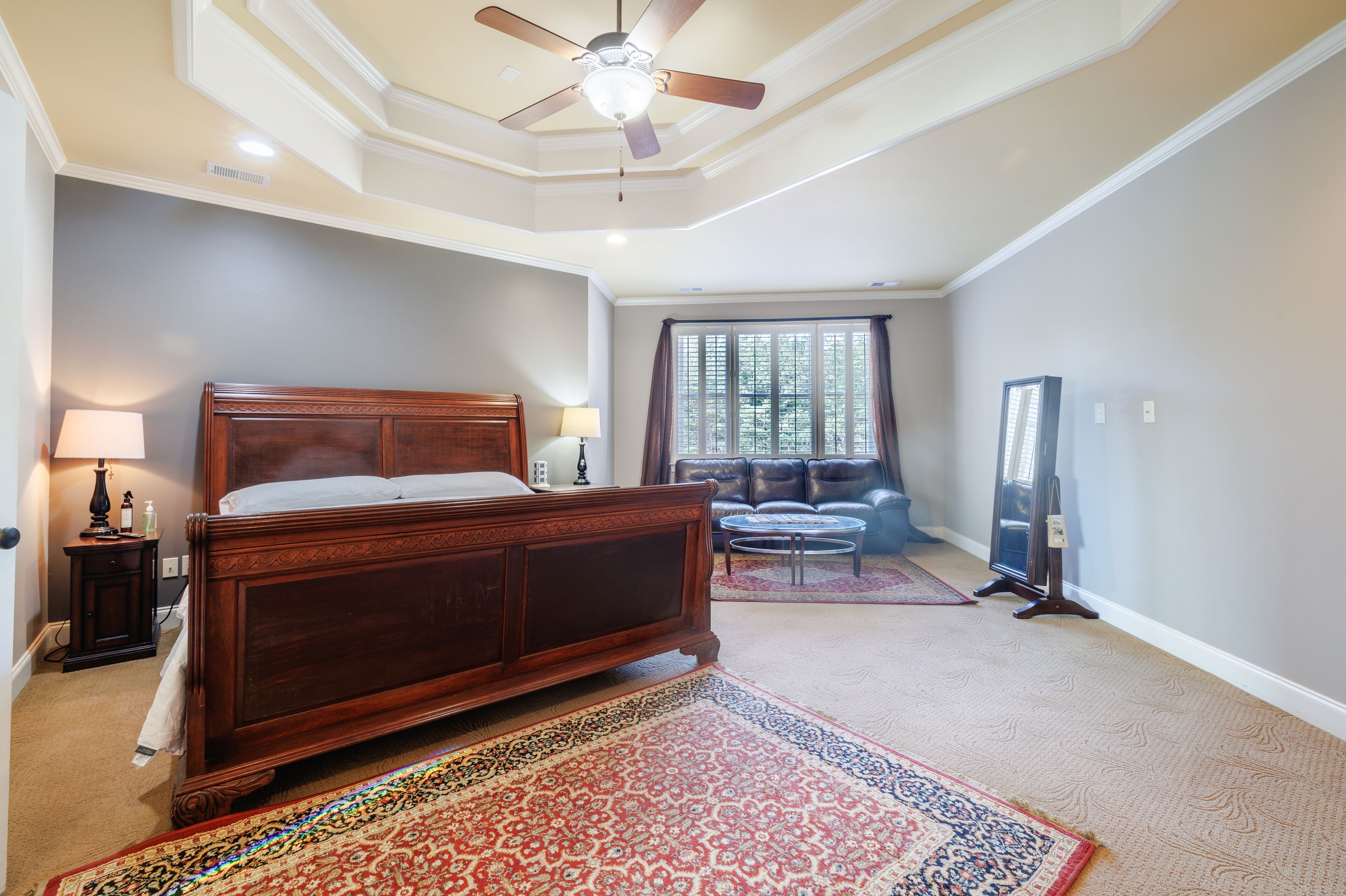 A bedroom with a wooden bed, a ceiling fan, a window with curtains, a sofa, and a mirror.