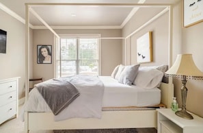 A bedroom with a bed, bedside table, lamp, and a window with blinds. - Luxury Estate•Saltwater Pool, Theater & Sleeps 16 (Lawrenceville)