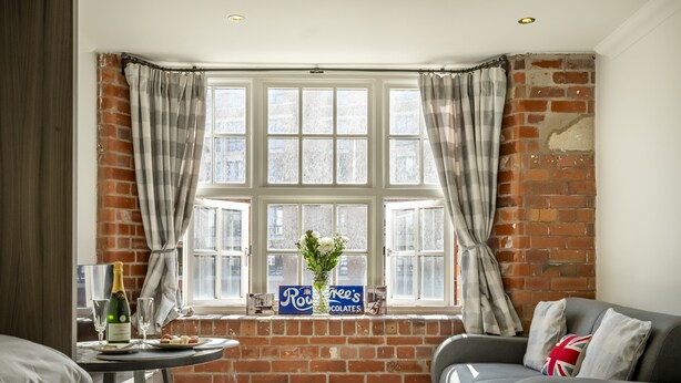 A hotel room with a brick wall, a large window with curtains, a table with a bottle and glasses, and a sofa with a pillow. - Cocoa Suites 35 - York city centre studio (with parking) - sleeps 3 (York)