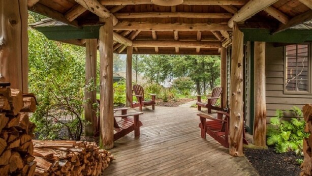 A wooden deck with chairs and a pile of firewood, surrounded by greenery.