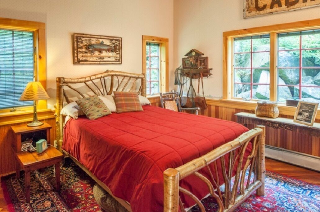 Una cama con colcha roja, cabecera de madera y mesita de noche con lámpara. Hay un cuadro en la pared y una ventana con vista a árboles.