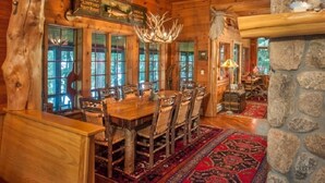 A rustic wooden dining area with a large table and chairs, a chandelier, and a stone wall.
