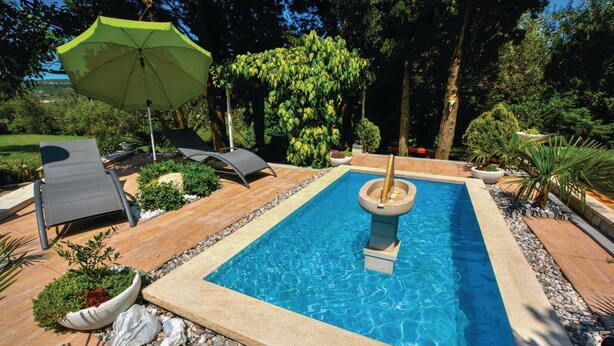 Una piscina rectangular con borde de piedra, rodeada por un lecho de guijarros. Hay dos sillones de descanso y una sombrilla.