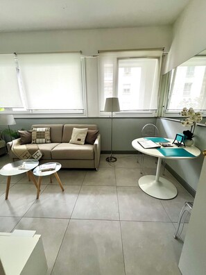 Ein modernes Wohnzimmer mit einer Couch, zwei Couchtischen mit Glasplatten, einem Beistelltisch, einer Lampe und Blick auf die Stadt durch große Fenster.