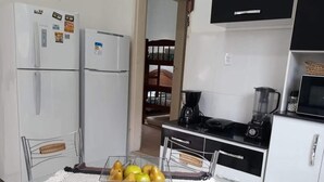 A kitchen with two white refrigerators, a black stove, and a microwave. There is a table with a woven placemat and a basket containing fruits. - Vacation Home in Arraial do Cabo (Praia Grande)