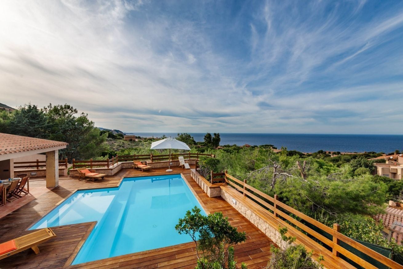 Piscina con cubierta de madera y sillones de descanso, rodeada de vegetación y con vistas al mar.