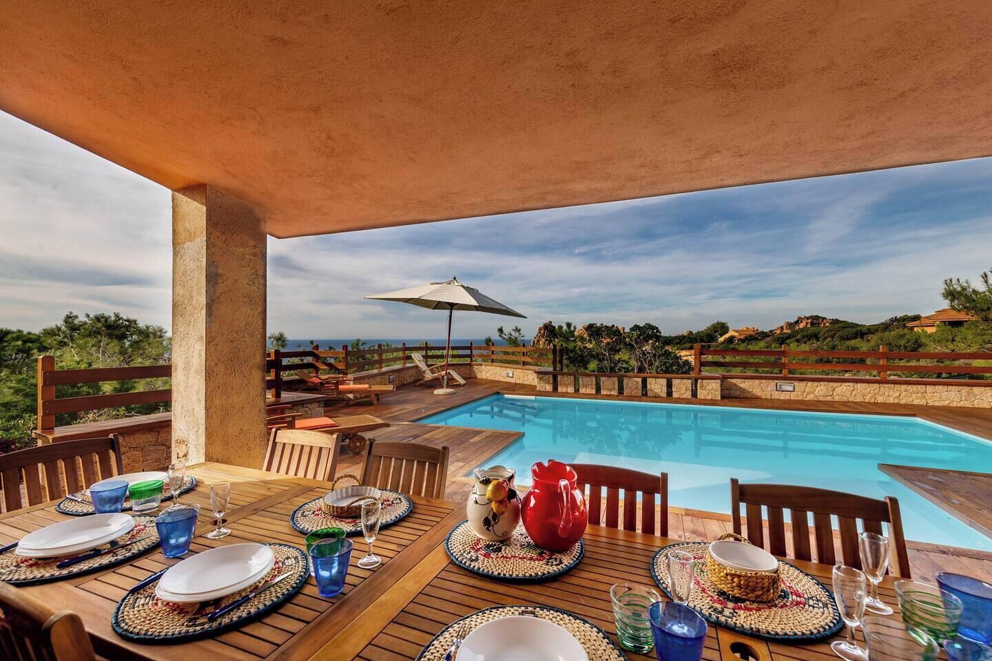 Un área de comedor junto a la piscina con mesas y sillas de madera, un paraguas grande y vistas al agua y al paisaje circundante.