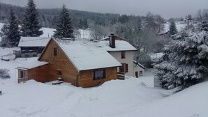A wooden cabin with a sloped roof, surrounded by snow-covered trees and a mountainous backdrop.