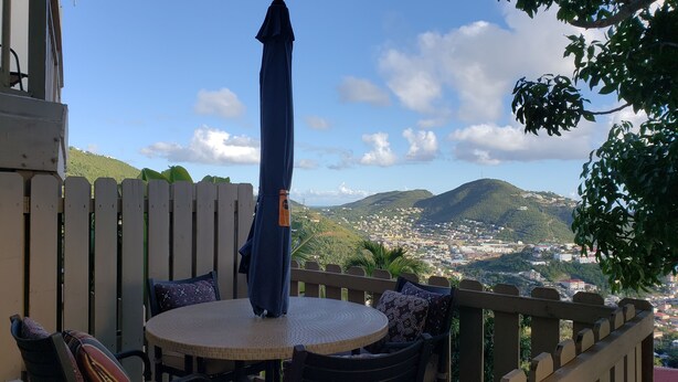 En balkon med et rundt bord og stole, en blå parasol, samt udsigt over en by og bakker i baggrunden.