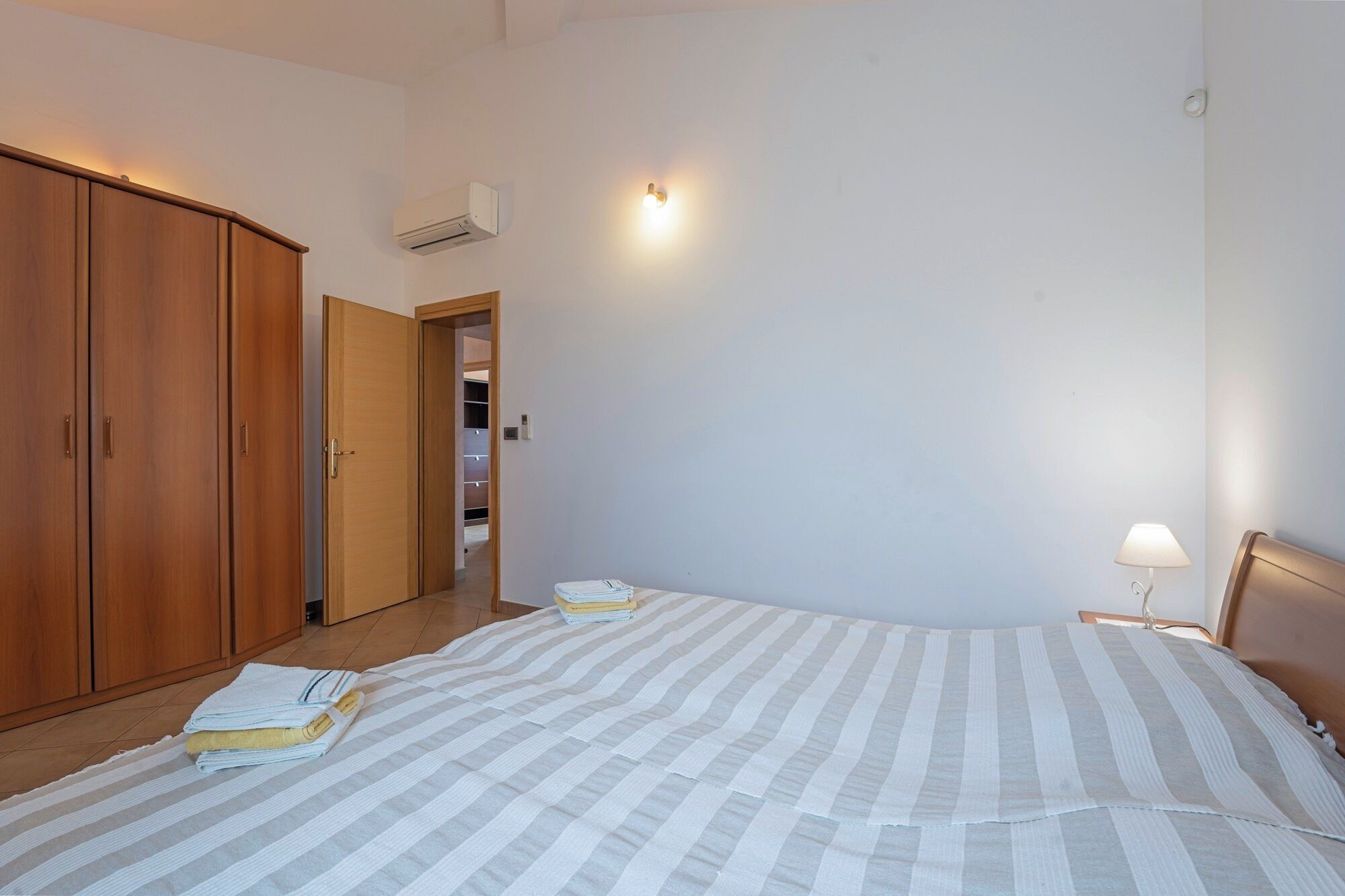 A bedroom with a bed, a wardrobe, an air conditioning unit, and a bedside lamp.