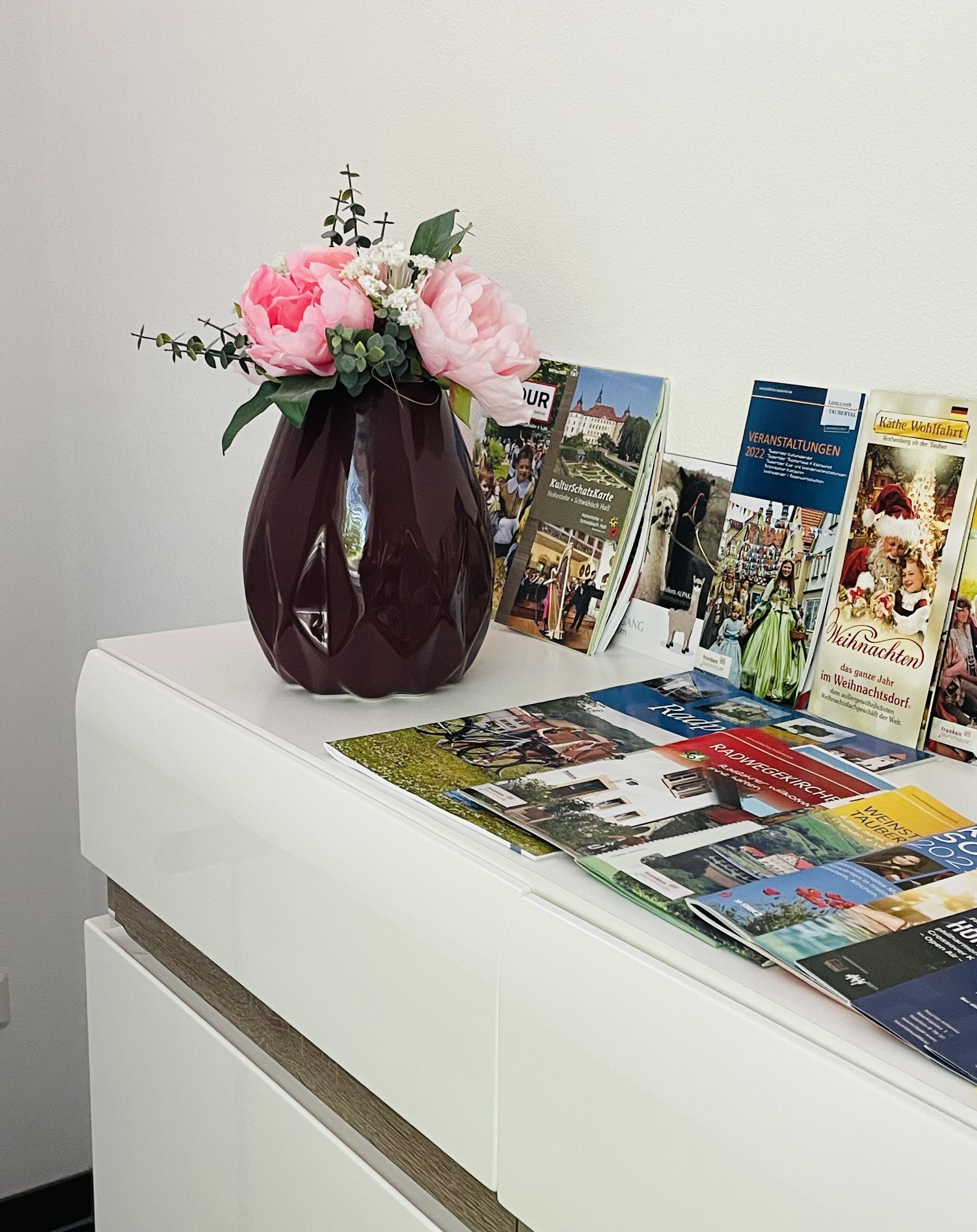 Ein weißer Tisch mit einer Vase voller pinkfarbener Blumen, Broschüren und einer Zeitschrift.