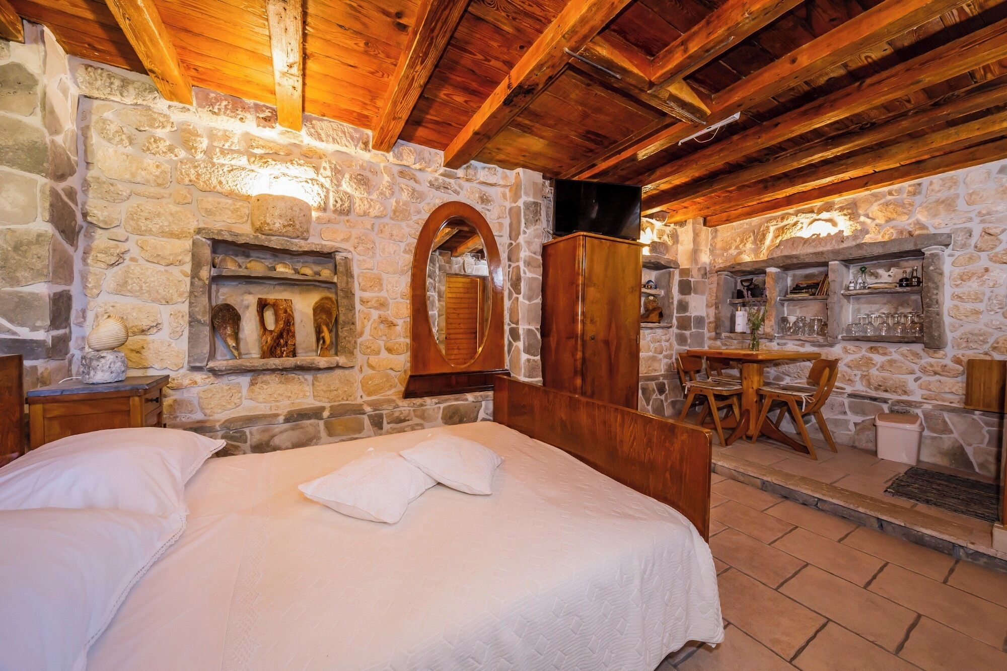 A cozy room with a stone wall, wooden ceiling beams, a bed with white linens, a wooden table with chairs, and a stone fireplace.