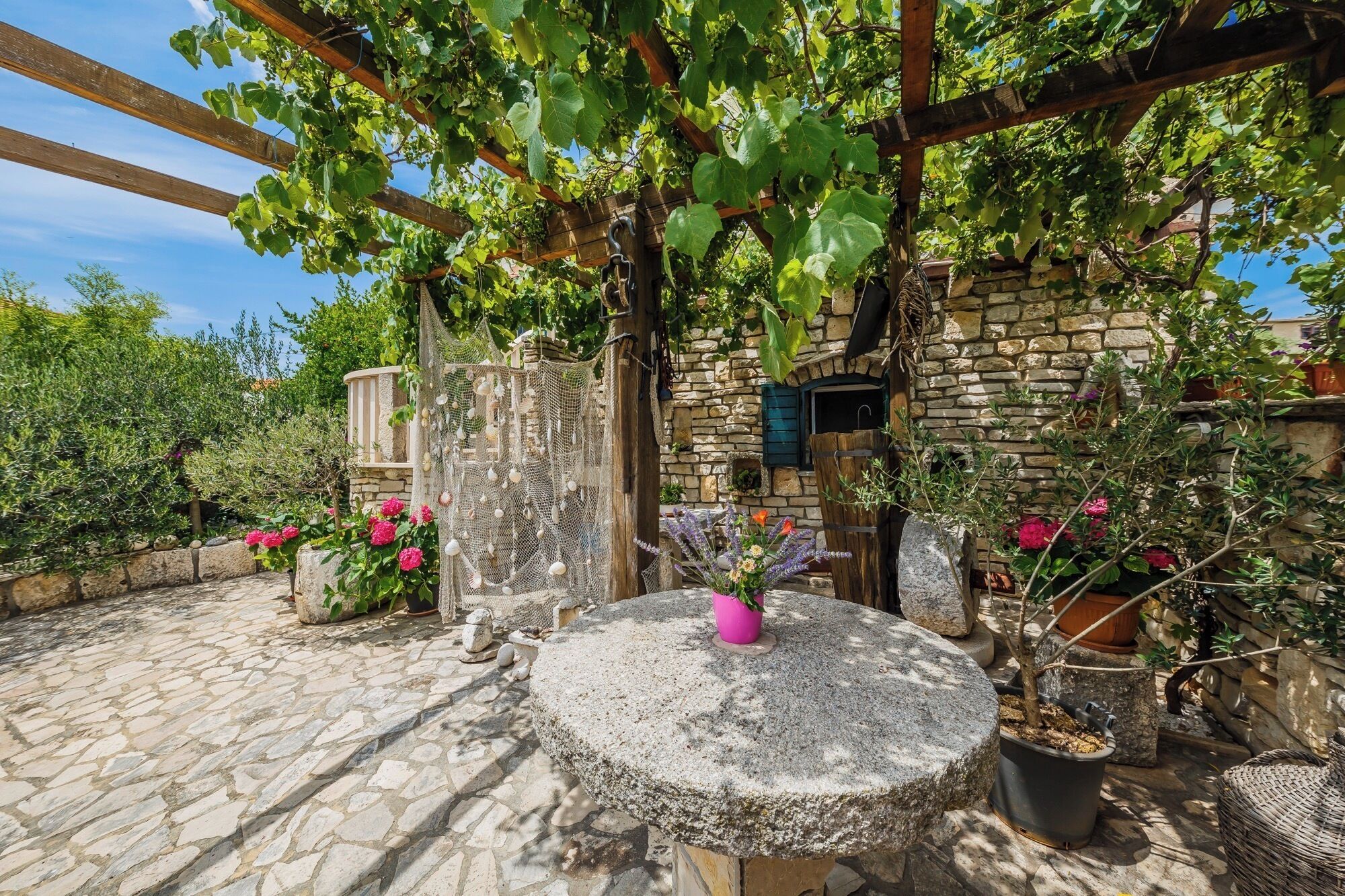 A stone building with a wooden pergola, potted plants, and a stone table with a pink vase.