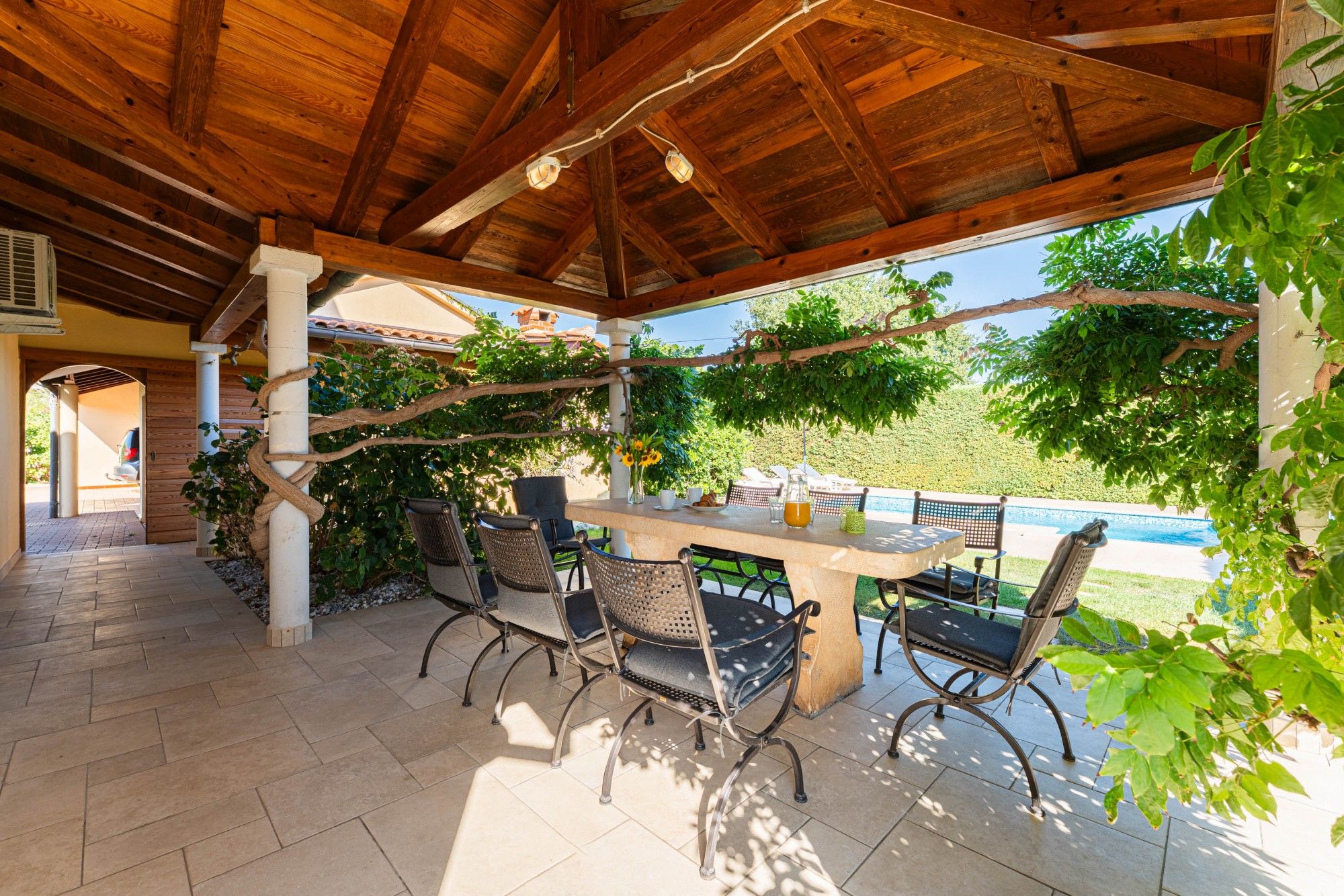 An outdoor patio with a wooden ceiling, a table with chairs, and a view of a swimming pool.