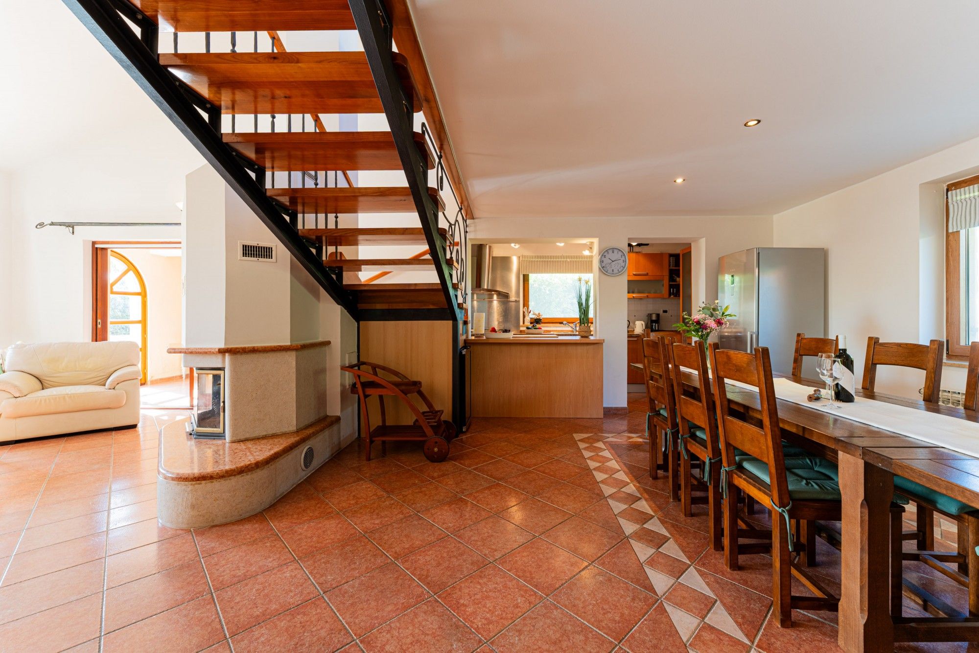 A spacious living area with a wooden staircase, a dining table with chairs, and a kitchen area with a refrigerator.