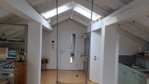 Un loft spacieux avec de hauts plafonds, une lucarne, un plancher en bois, une cuisine avec des armoires vertes et une porte visible donnant sur une autre pièce.