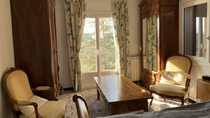 A room with a wooden bed, two chairs, a table, a mirror, and a window with floral curtains. - Beautifull villa, 3,360 sq.m garden; only 1 km away from the Saint-Endréol golf club (La Motte)
