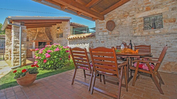 Un bâtiment en pierre avec une pergola en bois, une table avec des chaises et un pot de fleurs.