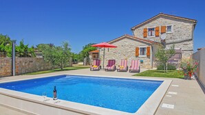 Une piscine entourée de chaises longues et agrémentée d’un parasol rouge, le tout bordé d’une maison en pierre avec un toit en tuiles.
