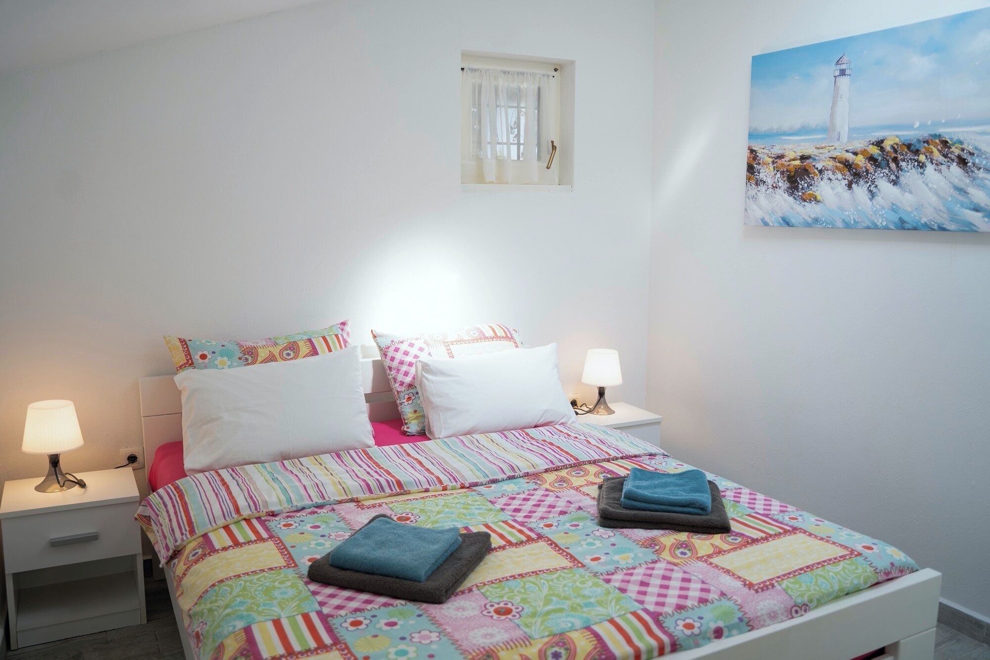 A bedroom with a bed, two bedside tables, a window with curtains, and a painting of a lighthouse.