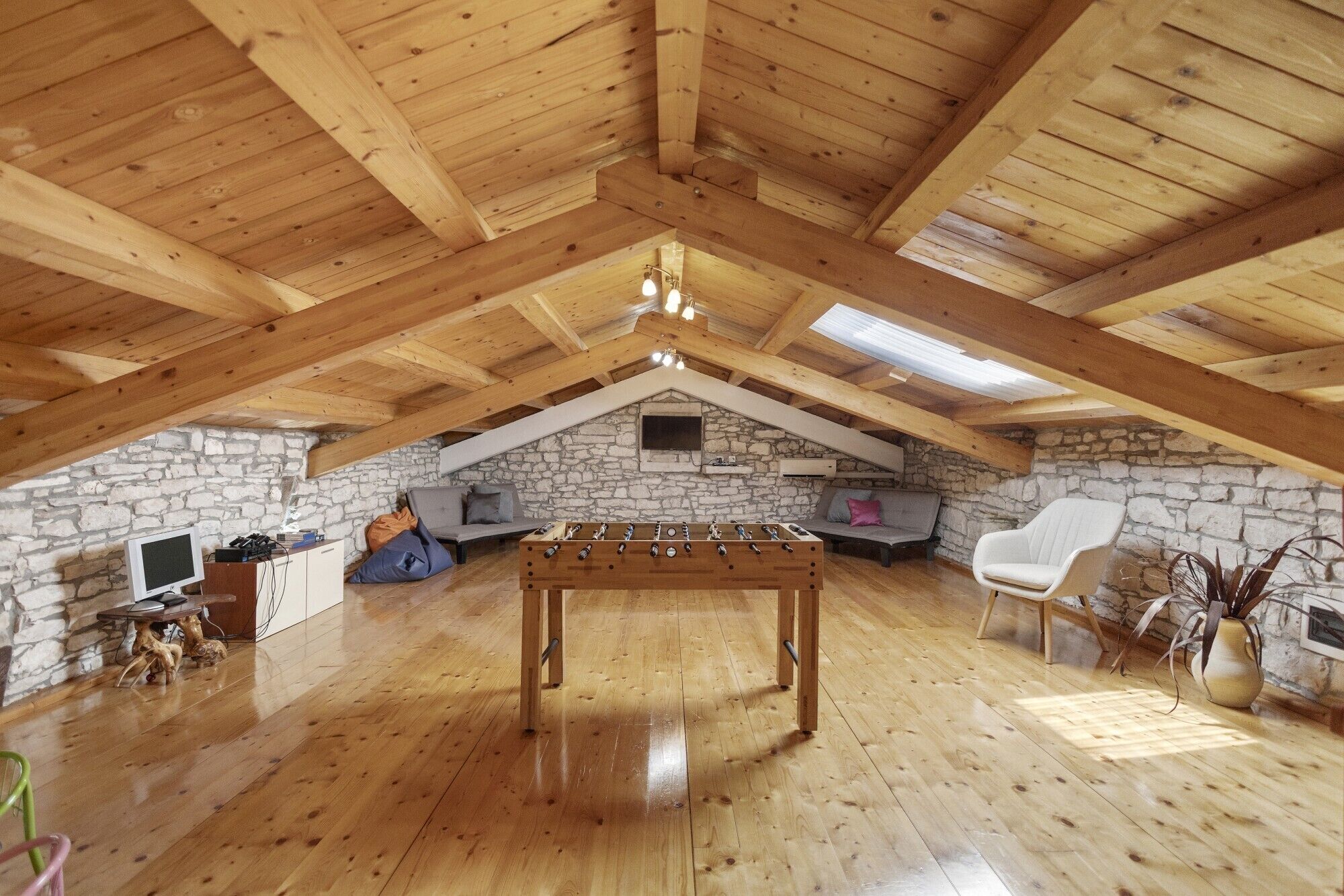 An attic room with a wooden ceiling, stone walls, a foosball table, a sofa, and a chair.