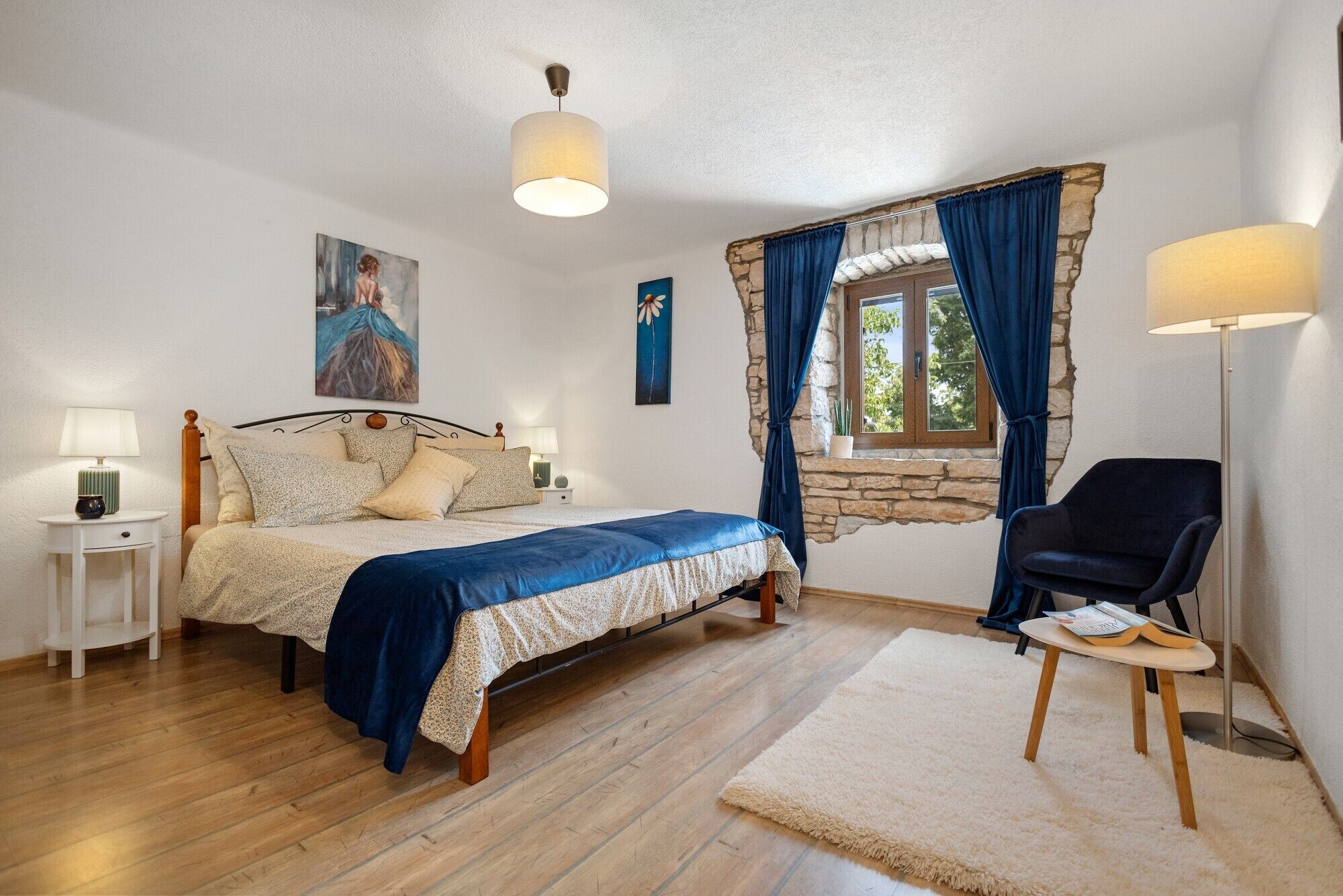 A bedroom with a bed, bedside tables, a chair, a window with curtains, and a painting on the wall.