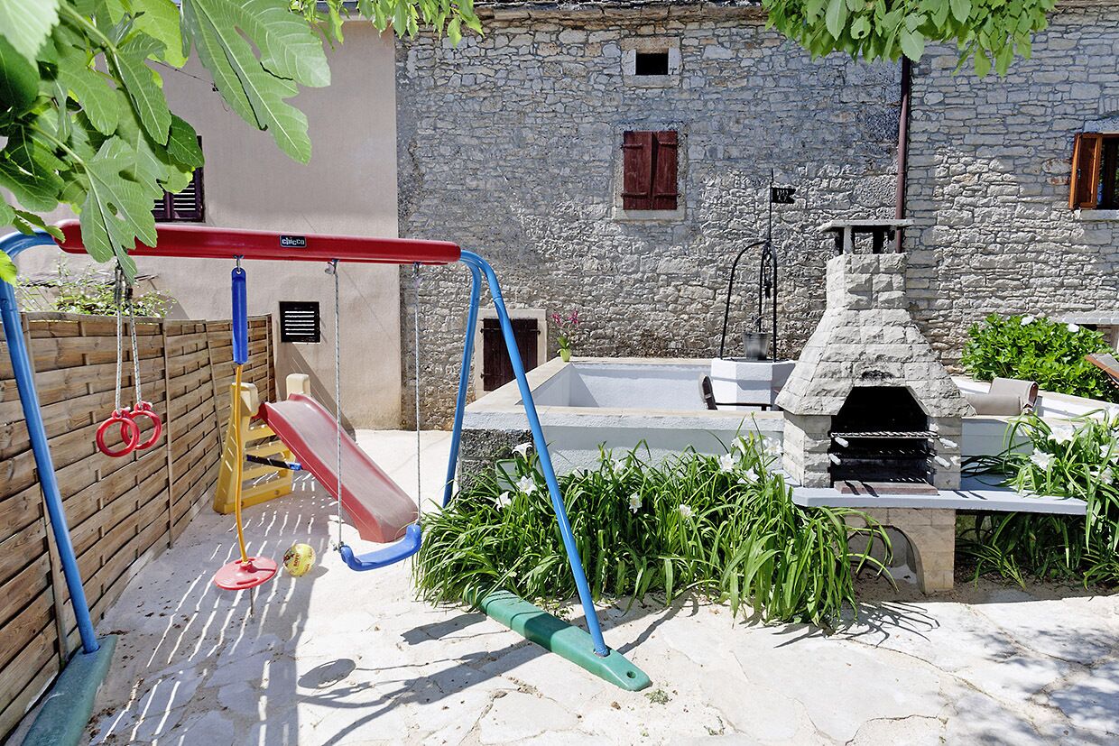 A children's playground with swings and a slide, a stone building with windows, and a stone barbecue grill.