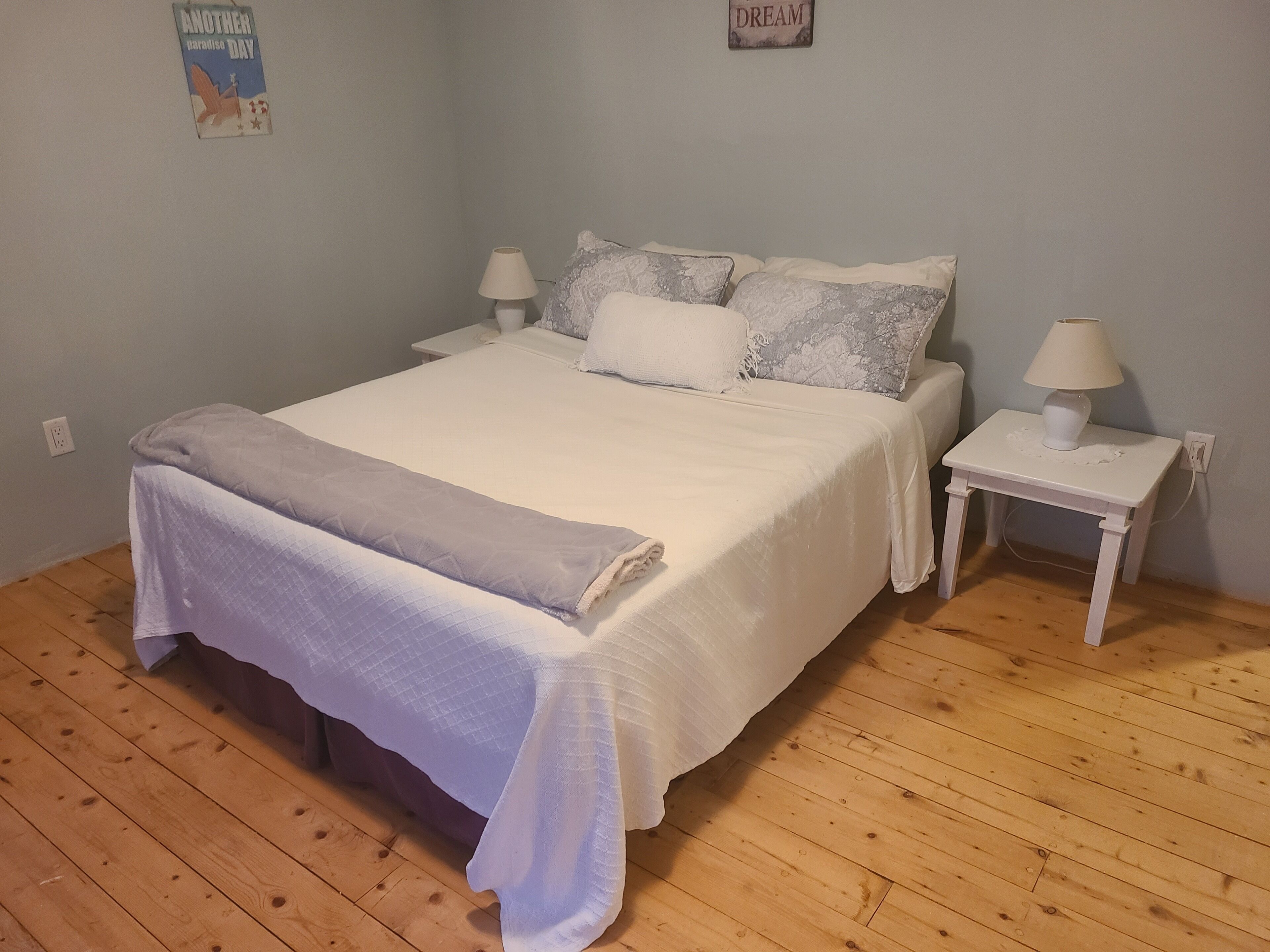 Una cama bien hecha con edredón blanco y manta gris, dos mesitas de noche con lámparas, y suelo de madera.
