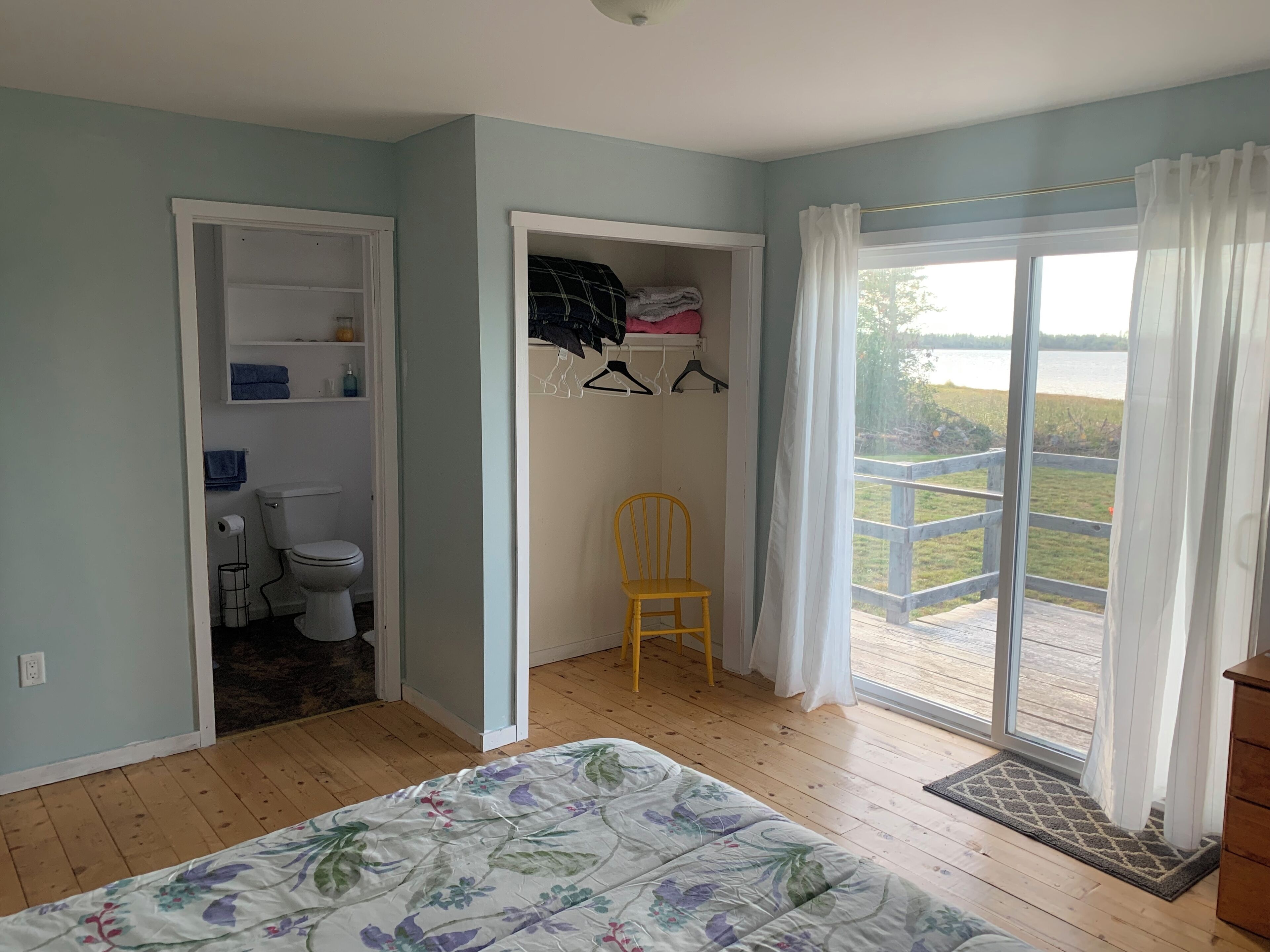 Un dormitorio con cama, un baño con inodoro, una silla y vistas a un lago a través de un ventanal grande.