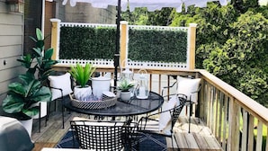 Une terrasse avec une table ronde, des chaises et un parasol blanc. On aperçoit, à l’arrière-plan, des plantes en pot et une clôture blanche.
