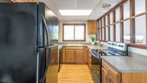 Une cuisine avec des meubles de rangement en bois, un réfrigérateur noir, une fenêtre avec des stores et un plan de travail avec une cuisinière.