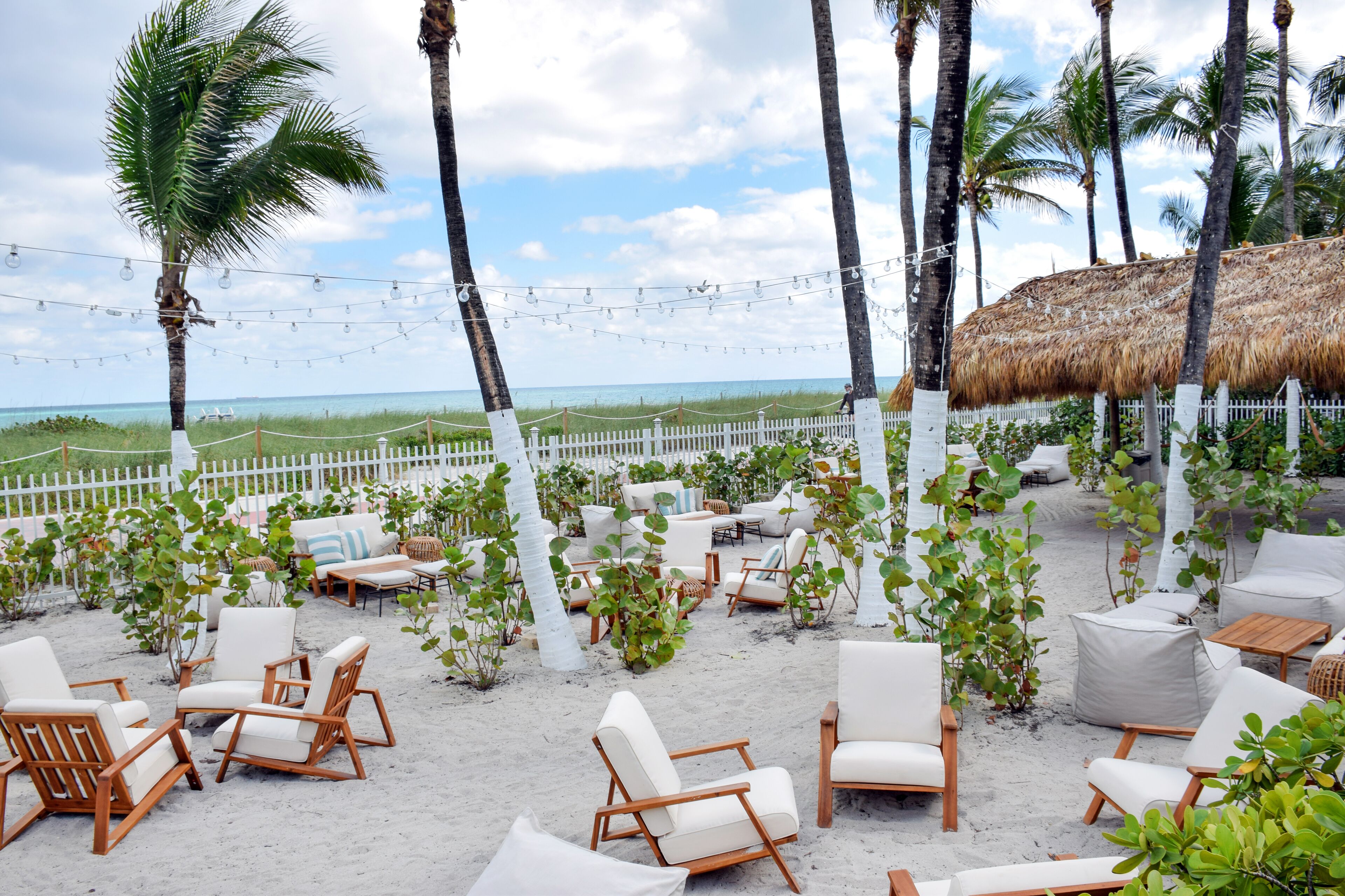 A beachside patio with white and wooden lounge chairs, a thatched-roof structure, and a white fence.