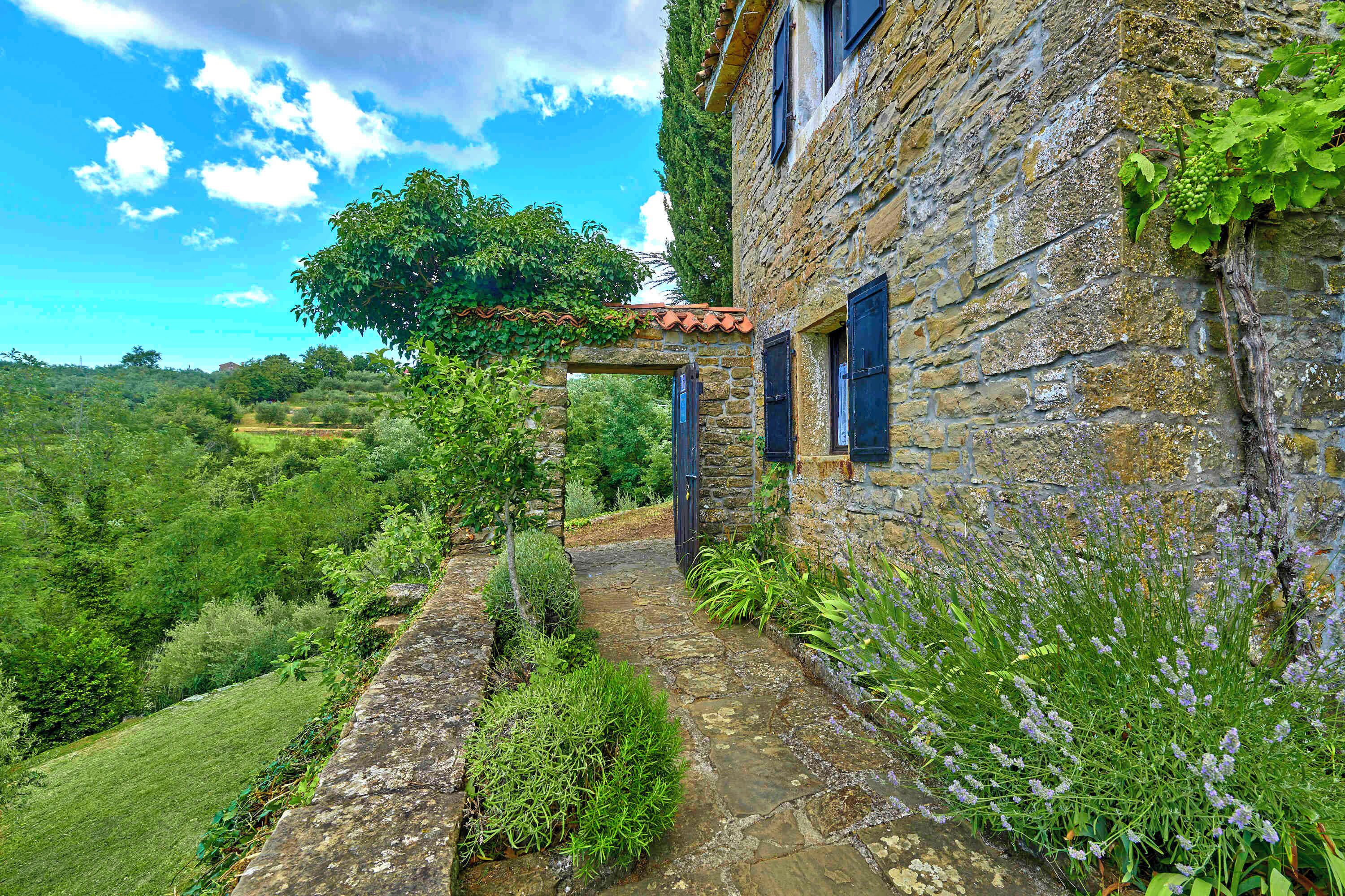 A stone building with a stone pathway leading to a wooden door, surrounded by lush greenery and a clear blue sky.