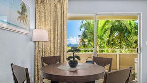 A round dining table with a potted plant, surrounded by chairs, in a room with a view of palm trees and a balcony.