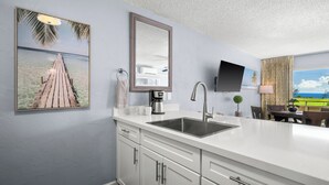 A modern kitchen with a white countertop, stainless steel sink, and a large mirror. There is a painting of a beach scene on the wall, a flat-screen TV mounted above the sink, and a dining area with a table and chairs visible in the background.