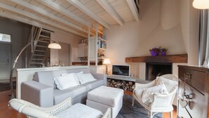 Ein Wohnzimmer mit einer Couch, einem Hocker, Sesseln, einem Kamin, einem Fernseher und einem Bücherregal.