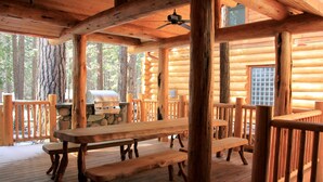 Una cabaña de madera con terraza techada, mesas de picnic y una parrilla.