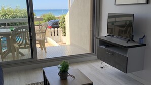 Una sala de estar moderna con un gran ventanal corredizo que da a un balcón con vistas al mar. Hay una mesita con una planta, un televisor sobre un mueble bajo y un escritorio con una silla.