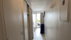 Un pasillo con una puerta a la izquierda, un radiador a la derecha y vistas al exterior por una puerta abierta.