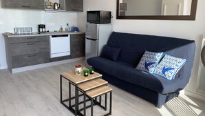 Una cocina moderna con armarios grises, un microondas y una cafetera. Hay un sofá azul oscuro con cojines estampados y una pequeña mesa de madera con una taza y vasos.