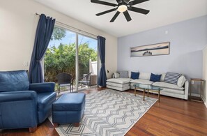 A modern living room with a sofa, ottoman, and coffee table, a ceiling fan, and a view through a glass door to an outdoor area.